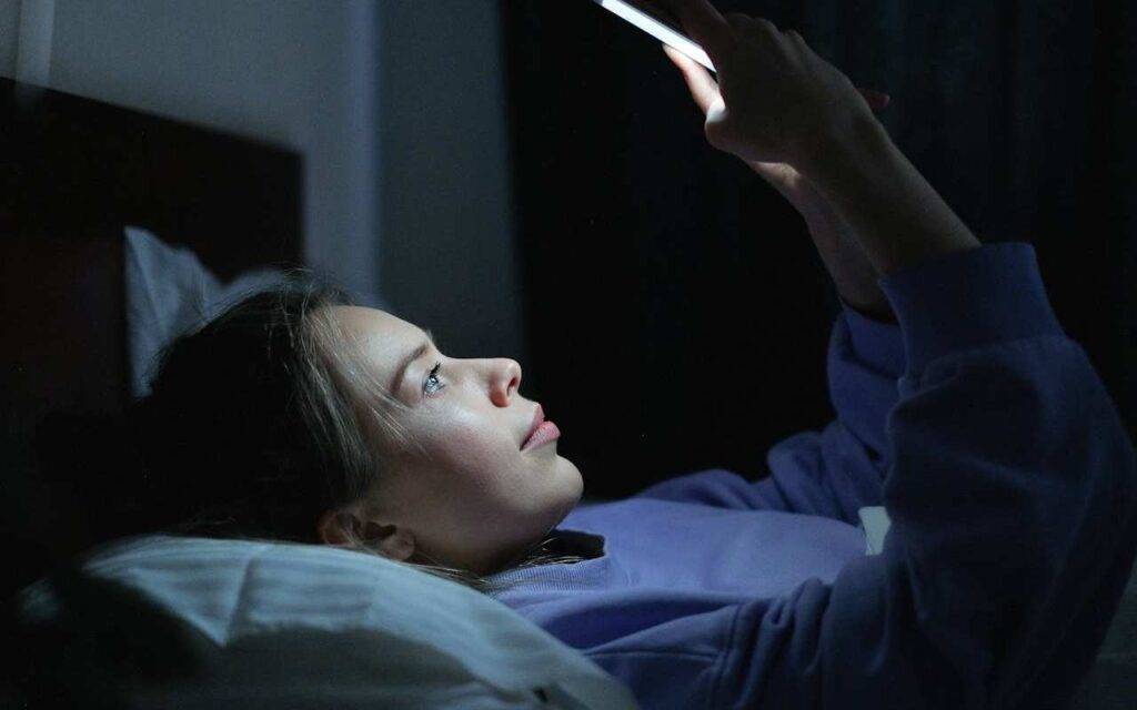 Devojka leži u krevetu i gleda u telefon.
