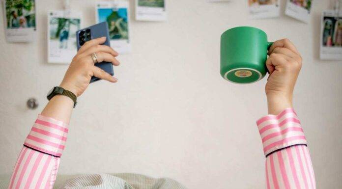 Devojka leži u krevetu pokrivena preko glave i drži u rukama telefon i šolju.