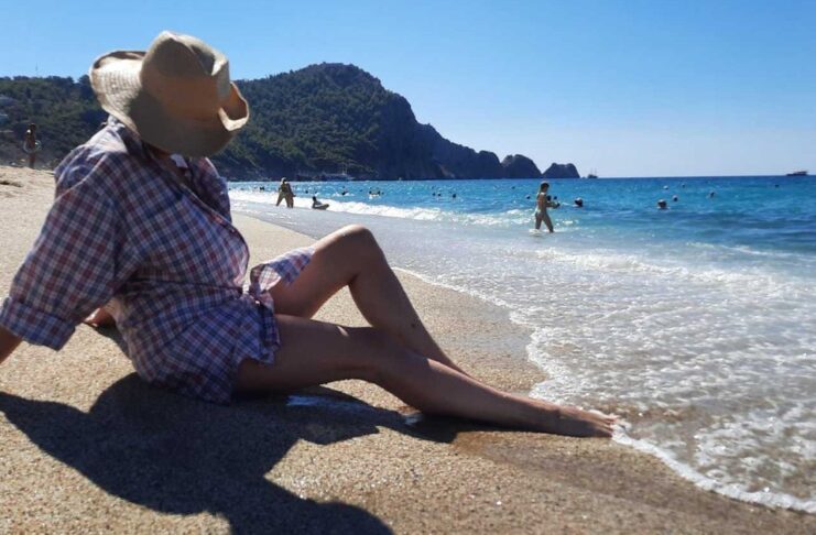 kreme za sunčanje, Žena na plaži sa šeširom u hladu, simbolizuje ljetnu zaštitu od sunca
