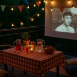 Ljetno kino na balkonu s lampicama, bokalom s voćem, jastučićima i projekcijom filma u toploj večeri