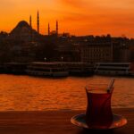 Istanbul grad kojem se vraćamo, Čaša turskog čaja na drvenom stolu uz zalazak sunca nad Bosforom, s pogledom na istanbulske džamije u jesenjim bojama.