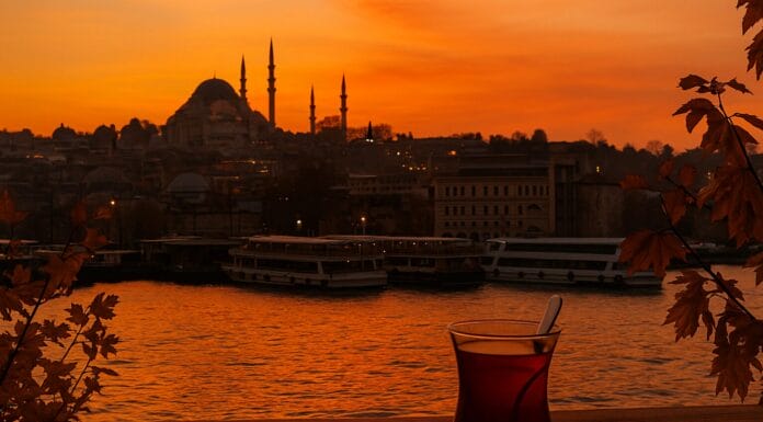 Istanbul grad kojem se vraćamo, Čaša turskog čaja na drvenom stolu uz zalazak sunca nad Bosforom, s pogledom na istanbulske džamije u jesenjim bojama.