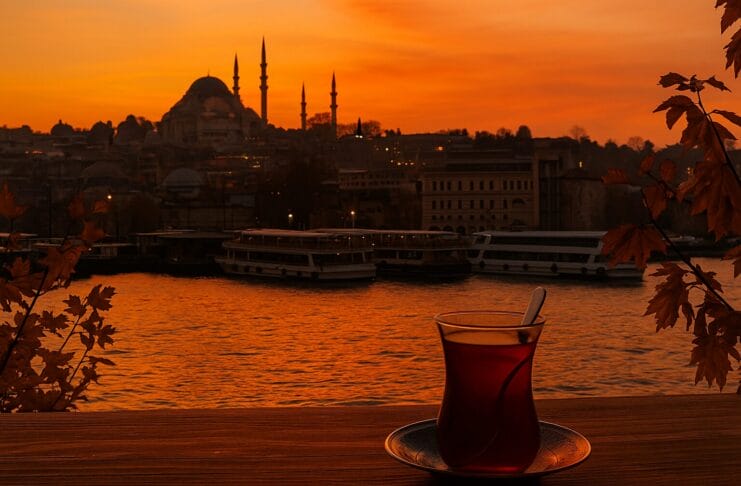 Istanbul grad kojem se vraćamo, Čaša turskog čaja na drvenom stolu uz zalazak sunca nad Bosforom, s pogledom na istanbulske džamije u jesenjim bojama.