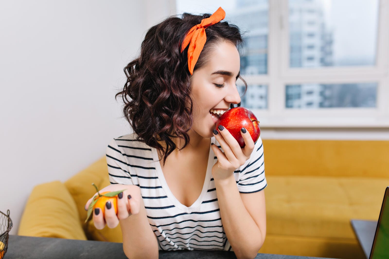 Portrait joyful excited young woman with curly cut hair enjoying red apple in modern apartment. Smiling, having fun, chilling at home, cosiness, relax, happiness.