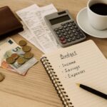 Svjetski dan štednje: Da li zaista znate gdje odlazi vaš novac? Međunarodni dan štednje – sto od svijetlog drveta s bilježnicom, računima, novčanikom, kovanicama i šoljicom kafe u toplom svjetlu, simbol planiranja troškova i štednje.