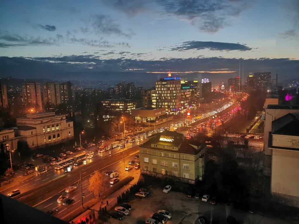 Ljubav u Sarajevu, Panorama Marijinog Dvora u Sarajevu u zimskom sutonu, sa gradskim svjetlima, automobilima i blagom večernjom maglom iznad grada.