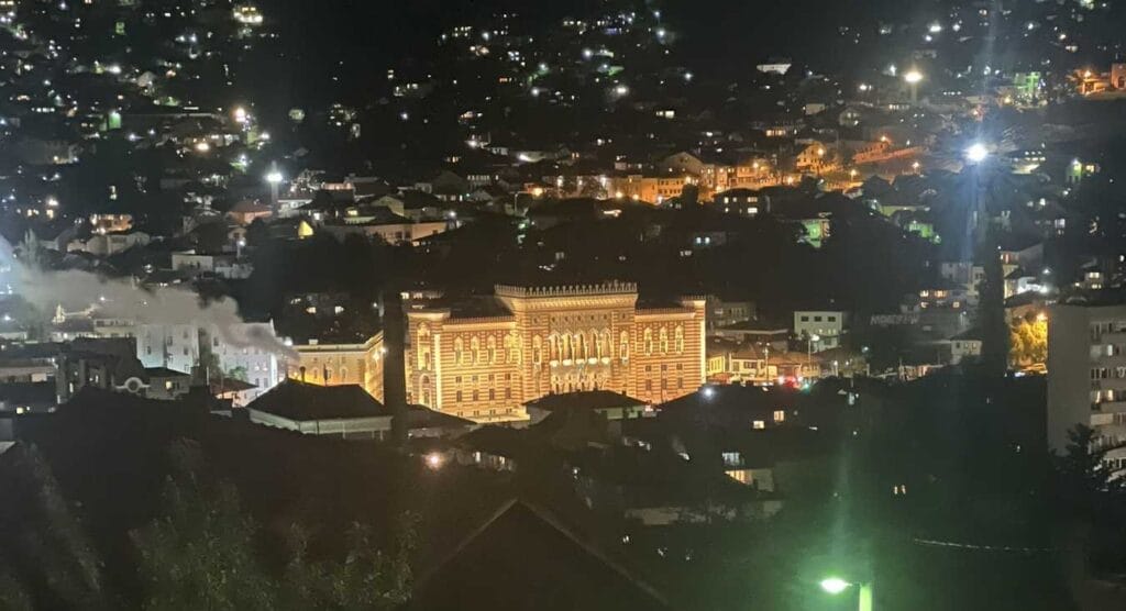 Ljubav u Sarajevu, Noćni pogled na sarajevsku Vijećnicu i okolne mahale, osvijetljene toplim žutim svjetlima pod tamnim nebom.